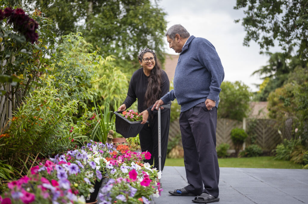 On older man with a mid skin tone and using a walking stick is talking a woman with a mid skin tone and long dark hair. She is smiling at the man and is holding a tub of flowers, which he is pointing at. They are in a garden, with lots of greenery, trees and colourful flowers. She seems to be placing the tub of flowers and maybe he is telling her what to do.