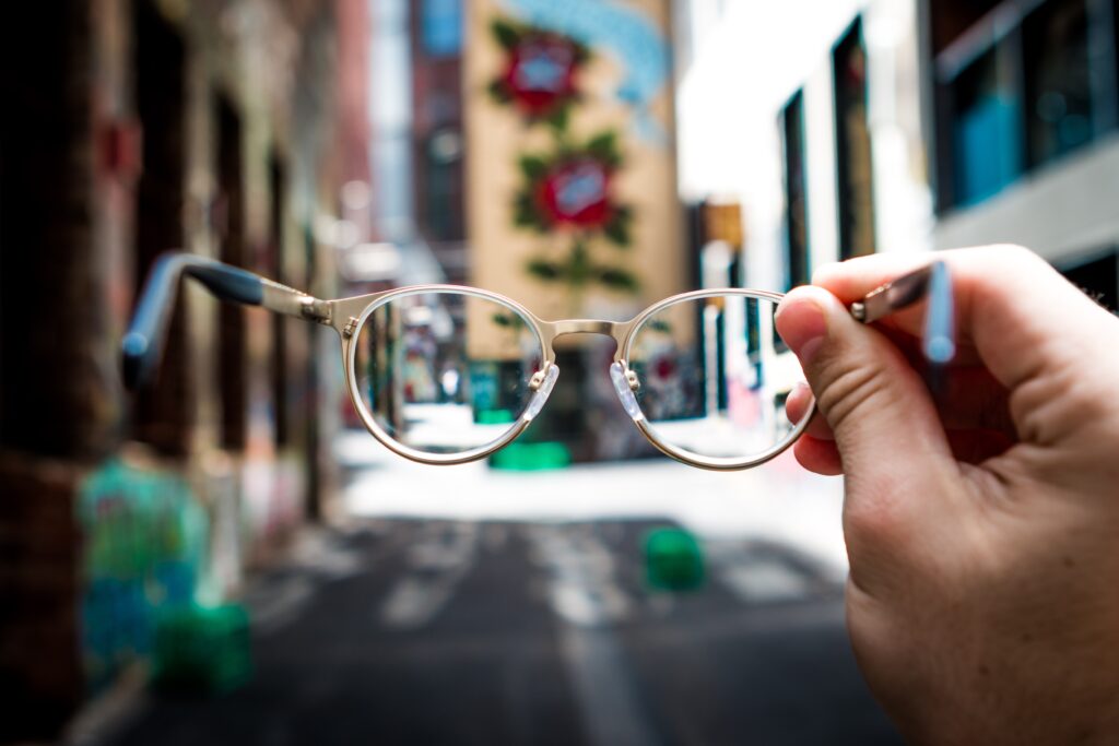 A pair of glasses is being held up to a street scene for the viewer to look through, The street scene is blurry around the glasses and a bit clearer through the lenses. The glasses and the hand holding them are closest to the viewer and in focus. It's a right hand with a light skin tone and the glasses have round gold frames and dark ear pieces. The street scene has taller buildings on either side and what seems to be a mural of flowers on the side of the building opposite the viewer. There are no people in sight.