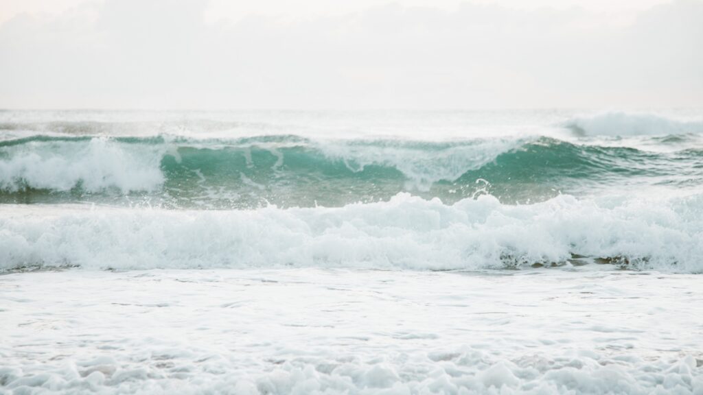 Surf's up! Waves are rolling towards the viewer, the sky is white, the sea is pale turquoise and the foam is white. It's easy to imagine the noise of the surf. No one is in sight.