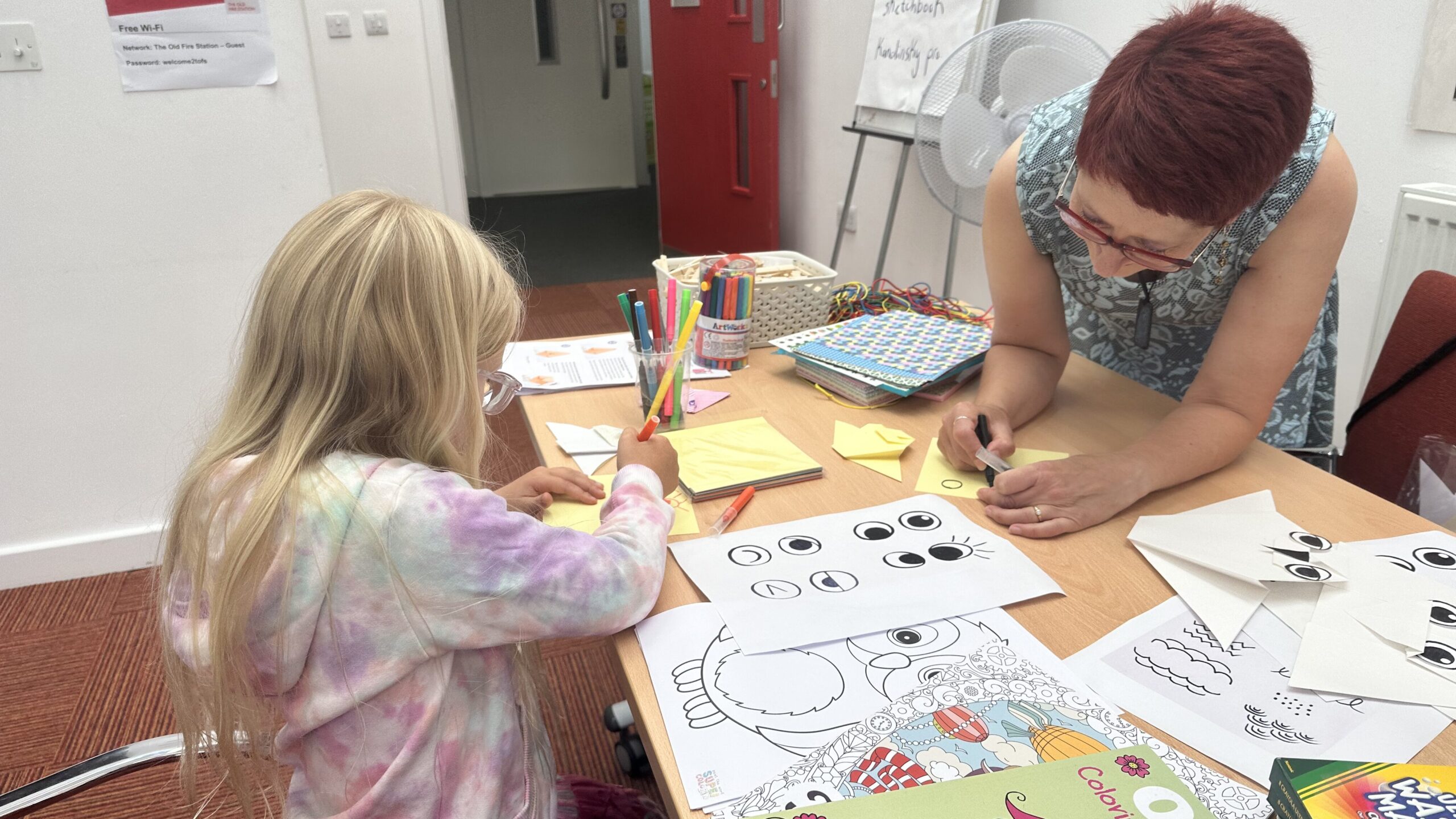 2 people are at a big table covered in origami paper, colouring pages, pens and paper owls. Jane, a woman with a light skin tone, short red hair and glasses is leaning over the table drawing with a black pen. A child with long blonde hair, a light skin tone, glasses and a top with lots of pastel colours is decorating her own paper owl.