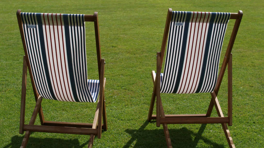 2 old-fashioned deckchairs are on a green lawn or in a park, their backs to the viewer. They have matching stipey cloth and wooden frames - the type that are tricky to put up unless you know how. The sun is shining, but no one is around to enjoy sitting in the chairs - yet!