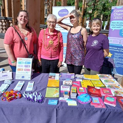 4 people stand behind a table covered with a purple cloth and lots of promotional items, including MindWell pens, bookmarks and rainbow wristbands and healthy living leaflets from Leeds Community Champions. In the background are tall, upright pieces of wood (a sculpture forest called Making a Stand) and the MindWell and Leeds Community Champions upright banners. The people all have light skin tones and are smiling and squinting into the sunshine. 1 has a pink top and long dark hair and wears a small ceremonial chain, the next has a pink jacket, short grey hair and glasses and the mayoral chains of office, then there’s a person in a multi-coloured sparkly top, with blonde hair fastened up and their hand shading their eyes. The final person wears a purple t-shirt and has short blonde hair.