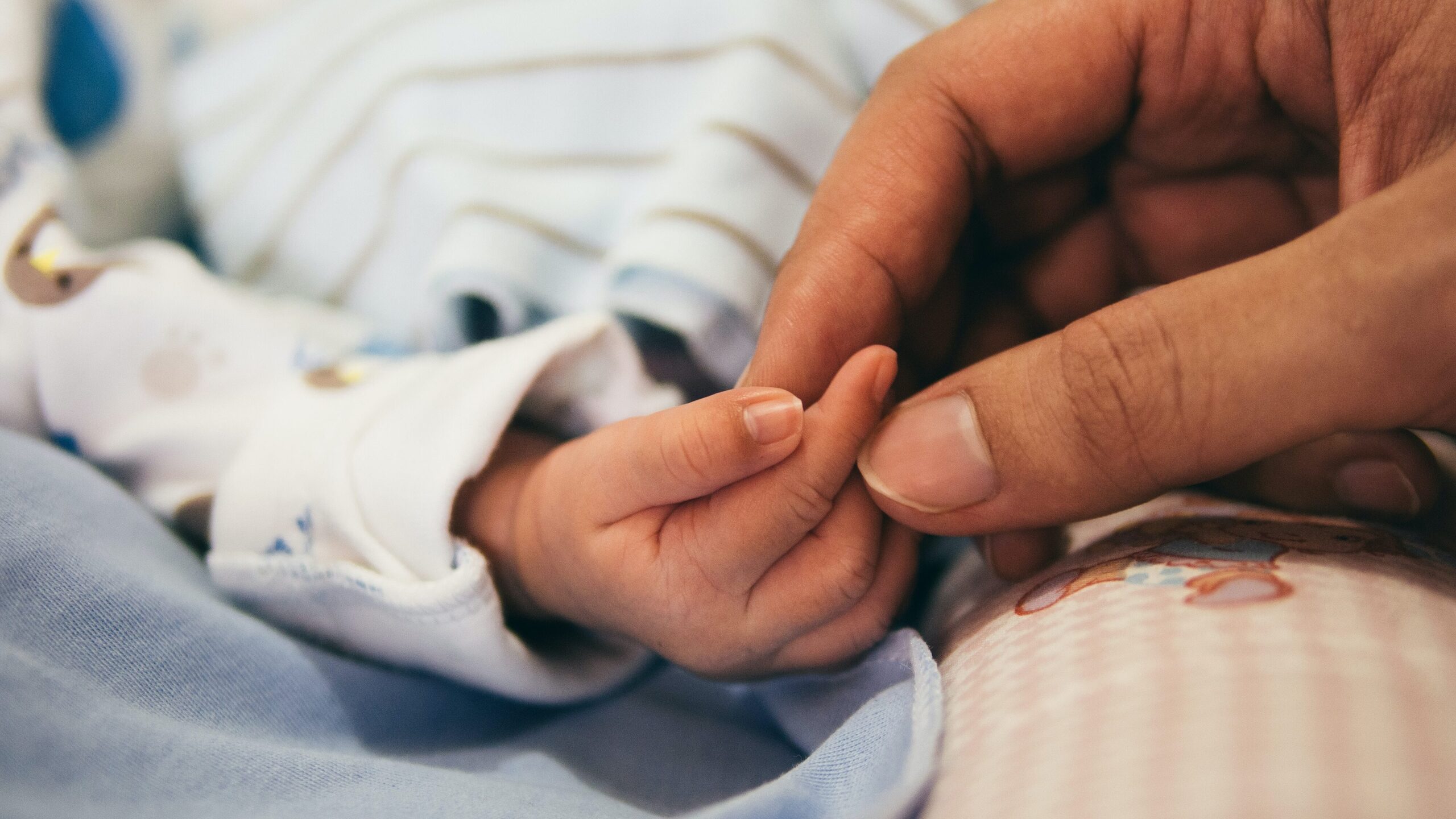 lose up of an adult hand holding the fingers of a small baby’s hand. Both people have a lighter skin tone, the baby is has a long white patterned sleeve and there are blankets in white, blue and peach.