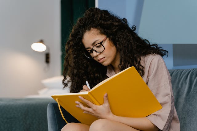 A person with a light to mid skin tone, long dark curly hair and wearing dark framed glassess and a beige shirt sits on a grey armchair . They lean over a large yellow journal as they write on the pages. There is a neatly made bed and wall lamp in the background.