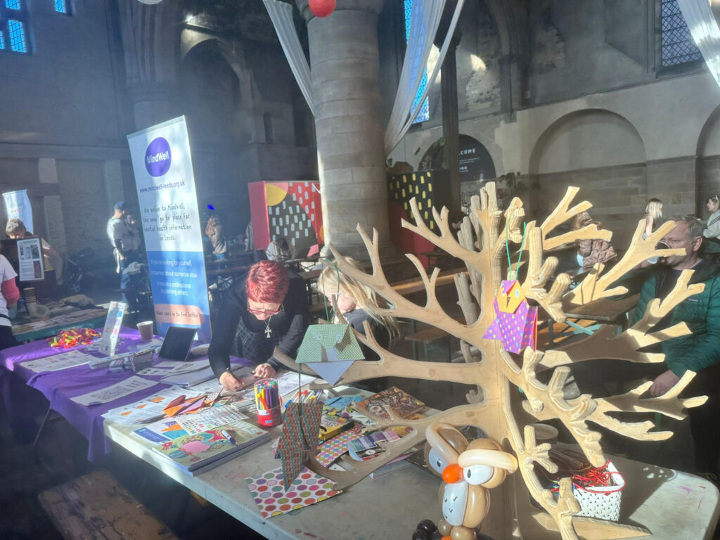 Jane, our workshop leader and a child are making owls at the MindWell stall at Kinder Leeds Festival. Jane has a light skin tone, short red hair and wears glasses and a black a grey jumper. The child has a pale sking tone and long, blonde hair. They are bent over the table as Jane demonstrates owl making.