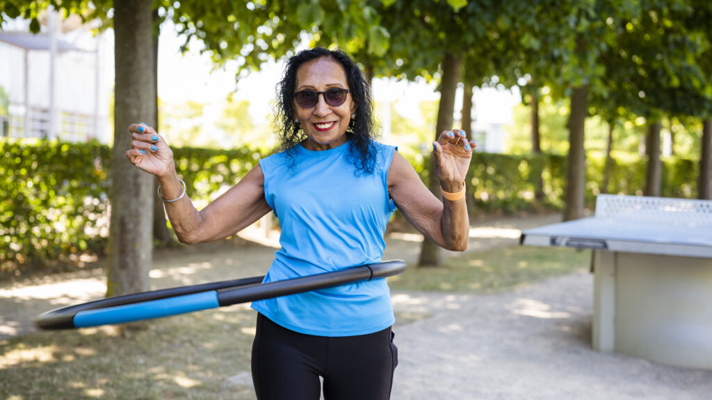 A person with a light to mid skin tone, shoulder length dark curly hair, dark glasses, a big smile and wearing a bright blue top is hula hooping in a leafy park.