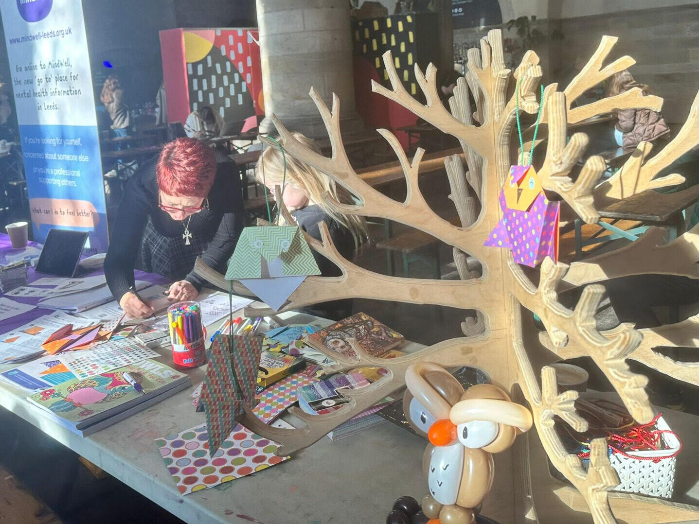 Jane, our workshop leader and a child are making owls at the MindWell stall at Kinder Leeds Festival. Jane has a light skin tone, short red hair and wears glasses and a black a grey jumper. The child has a pale sking tone and long, blonde hair. They are bent over the table as Jane demonstrates owl making.