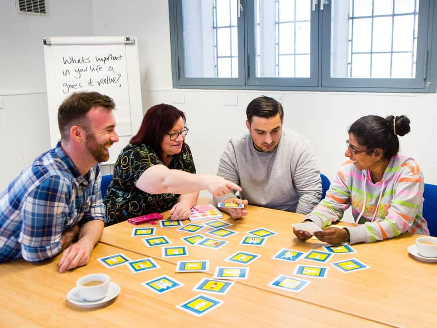 A meeting room with 4 people sitting around a table, sorting blue and yellow cards. They look focused or are smiling. A flipchart in the background says 'What is important in your life and gives value?' The photo is from a Leeds Recovery College news story.