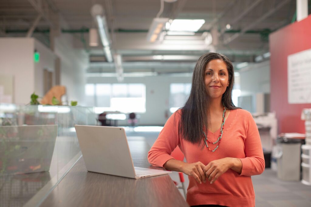 Confident woman in a bright, open-plan office, standing next to her laptop and smiling warmly.