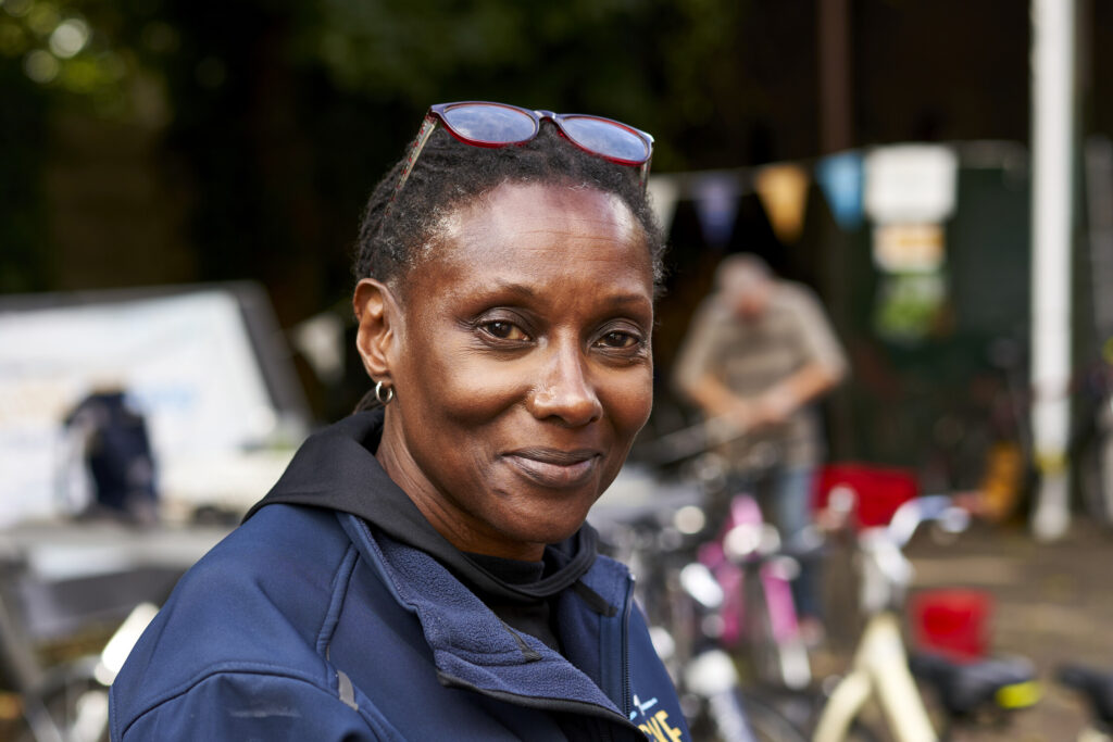 Smiling woman in a navy jacket with glasses on her head, standing outdoors with blurred bicycles and bunting in the background.