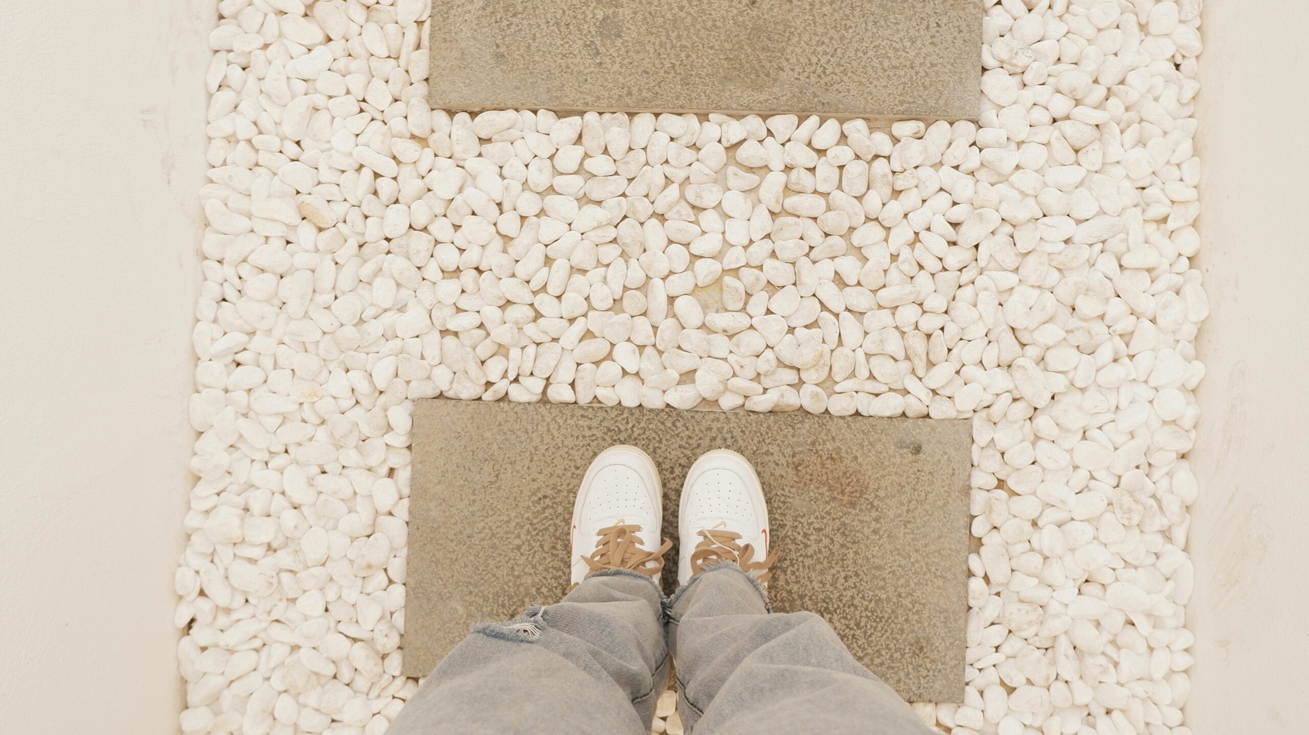 View looking down at a person standing on a stone paving slab surrounded by white pebbles, with another paving slab ahead.